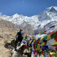 Uśmiechnięta turystka na tle majestatycznych szczytów Annapurna Base Camp, otoczona kolorowymi flagami modlitewnymi.