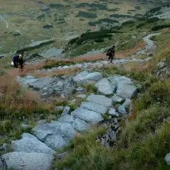 Dwóch turystów z plecakami idzie kamienistym szlakiem w górach.