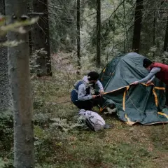 Dwóch turystów rozkłada zielony namiot w lesie. To ich patenty pod namiot, by nocleg był udany.