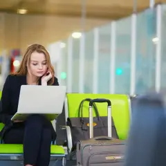 Kobieta w czarnej marynarce siedzi na zielonym krześle na lotnisku, pracując na laptopie. Obok niej walizka i torba. Zastanawia się, czy można anulować bilet lotniczy.