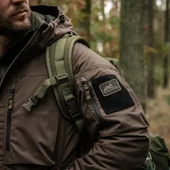 Mężczyzna w brązowej kurtce z naszywką Helikon-Tex, symbol polskie marki outdoorowe, w lesie.
