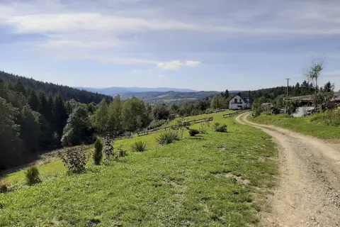 Droga wije się przez zielone wzgórza, prowadząc do domu na skraju lasu. Piękny widok na Beskidy, idealny na mały szlak beskidzki.