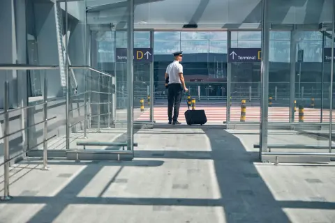 Pilot z walizką czeka przed wejściem do terminalu. Jak dolecieć na Azory? Kierunek Terminal D.