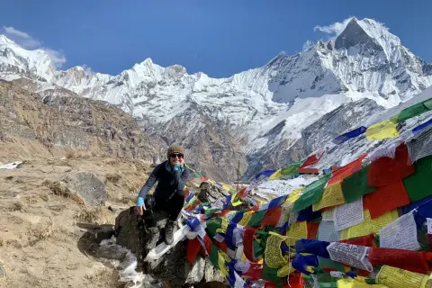 Uśmiechnięta turystka na tle majestatycznych szczytów Annapurna Base Camp, otoczona kolorowymi flagami modlitewnymi.