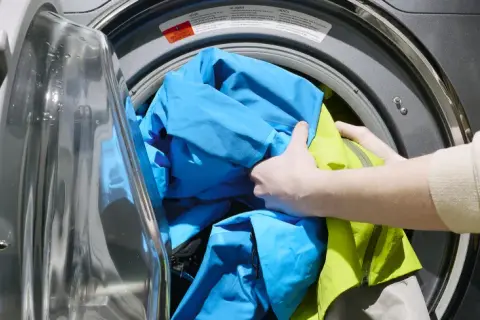 Ręce wkładają niebieską i zieloną kurtkę do pralki. Dowiedz się, jak prać Gore-Tex, aby zachować jego właściwości.