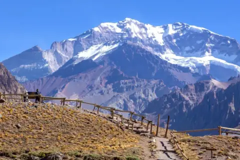 Widok na ośnieżone góry z Aconcagua, z kamienistym szlakiem i drewnianym ogrodzeniem na pierwszym planie.