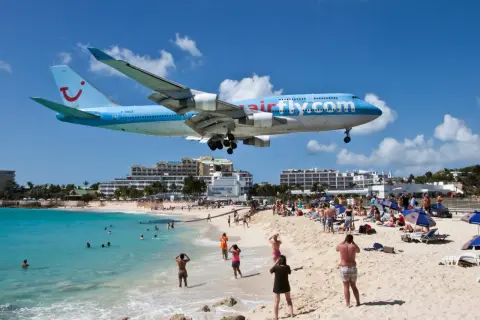 Samolot Airfly.com nisko przelatuje nad plażą pełną turystów. To słynne lotnisko przy plaży, gdzie można poczuć podmuchy odrzutowców.