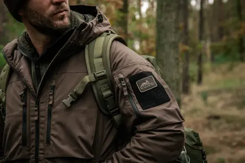 Mężczyzna w brązowej kurtce z naszywką Helikon-Tex, symbol polskie marki outdoorowe, w lesie.