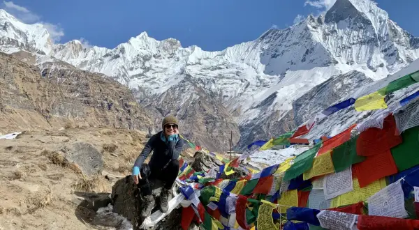 Uśmiechnięta turystka na tle majestatycznych szczytów Annapurna Base Camp, otoczona kolorowymi flagami modlitewnymi.