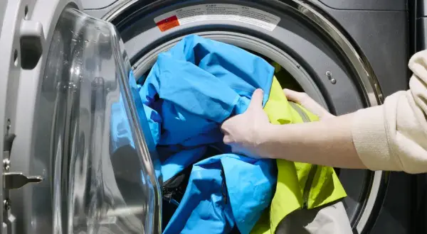 Ręce wkładają niebieską i zieloną kurtkę do pralki. Dowiedz się, jak prać Gore-Tex, aby zachować jego właściwości.