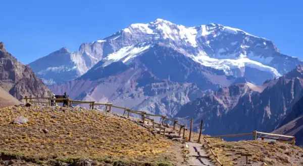 Widok na ośnieżone góry z Aconcagua, z kamienistym szlakiem i drewnianym ogrodzeniem na pierwszym planie.