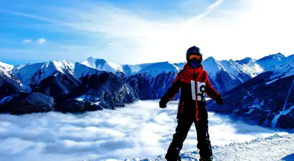 Snowboardzista w hardshellowej kurtce stoi na szczycie góry, nad morzem chmur.