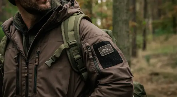 Mężczyzna w brązowej kurtce z naszywką Helikon-Tex, symbol polskie marki outdoorowe, w lesie.