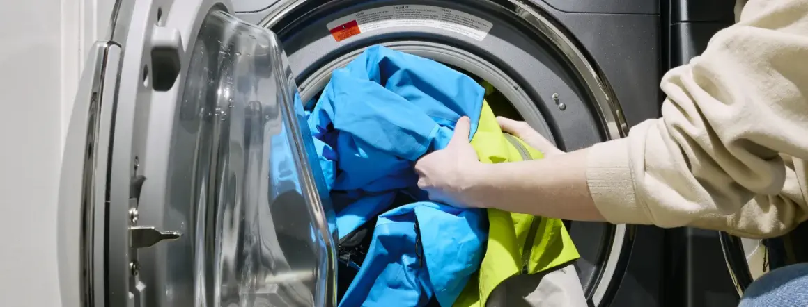 Ręce wkładają niebieską i zieloną kurtkę do pralki. Dowiedz się, jak prać Gore-Tex, aby zachować jego właściwości.