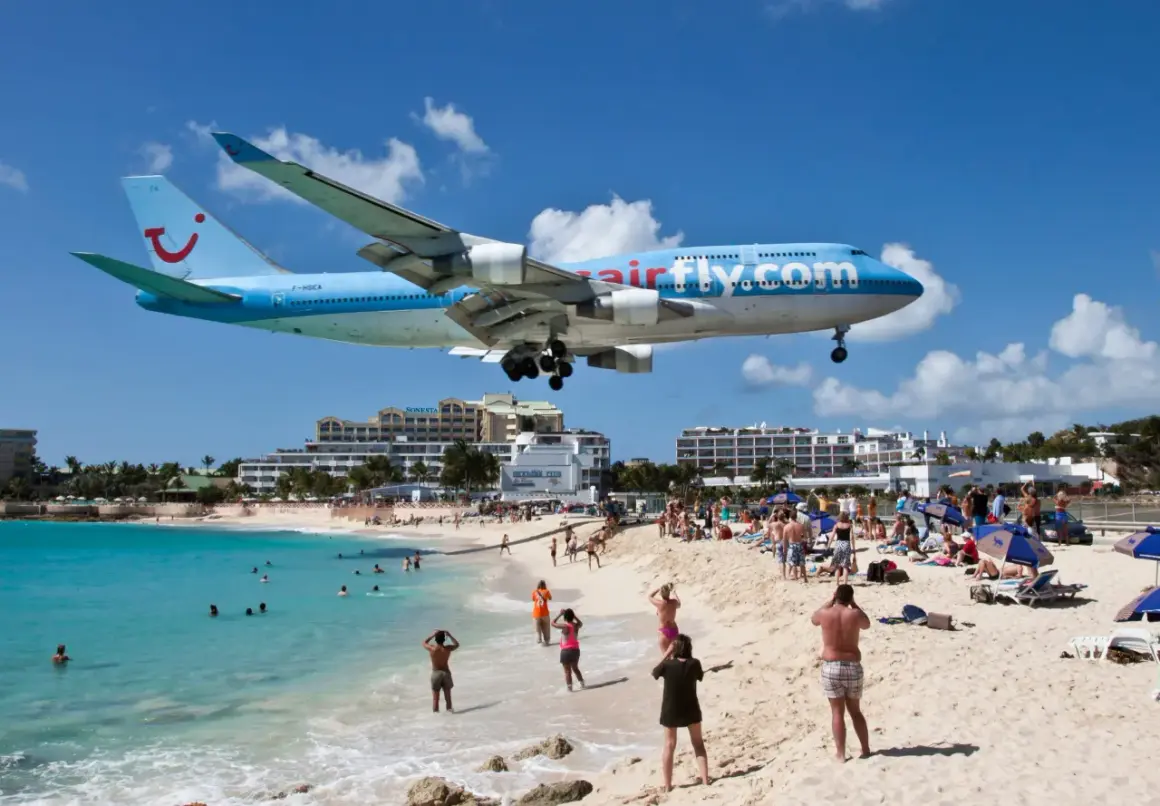 Samolot Airfly.com nisko przelatuje nad plażą pełną turystów. To słynne lotnisko przy plaży, gdzie można poczuć podmuchy odrzutowców.