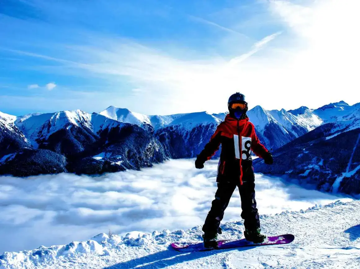 Snowboardzista w hardshellowej kurtce stoi na szczycie góry, nad morzem chmur.