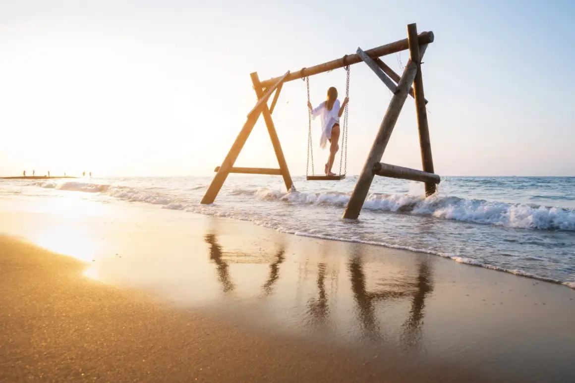 Dziewczyna na huśtawce na plaży w Turcji, fale obmywają piasek, złote światło zachodu słońca.