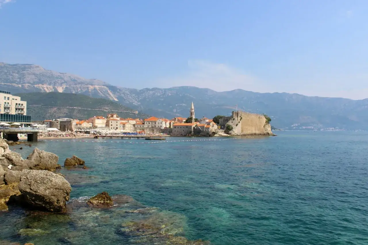 Budva co zobaczyć: Stare Miasto z cytadelą i kościołem na skalistym cyplu, plaża i g&oacute;ry w tle.