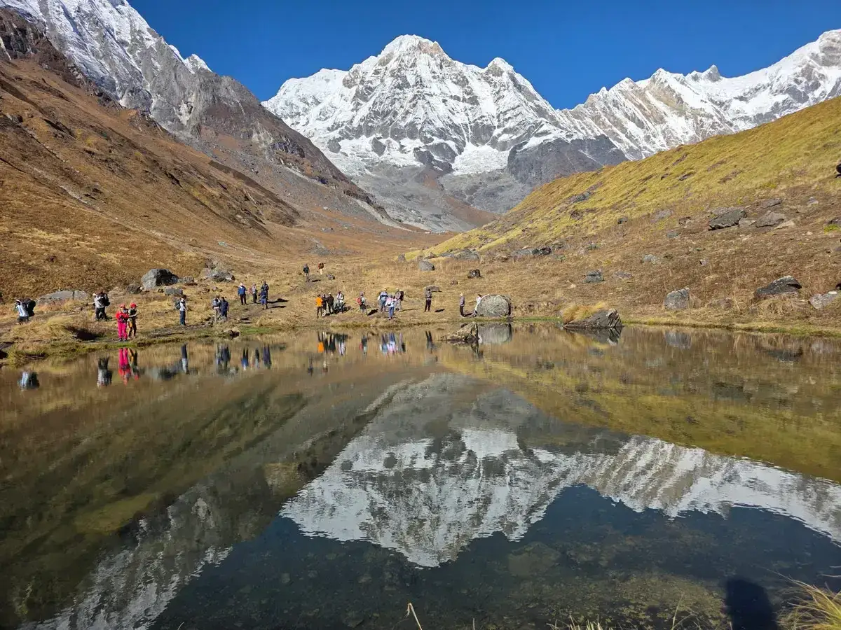 Grupa turystów podziwia odbicie ośnieżonych szczytów w spokojnej tafli jeziora. To widok z Annapurna Base Camp.