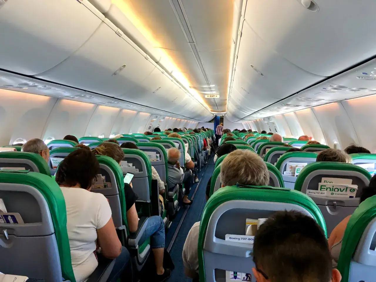 Wnętrze samolotu Transavia, pasażerowie siedzą w rzędach, bagaż podręczny schowany w schowkach nad głowami.