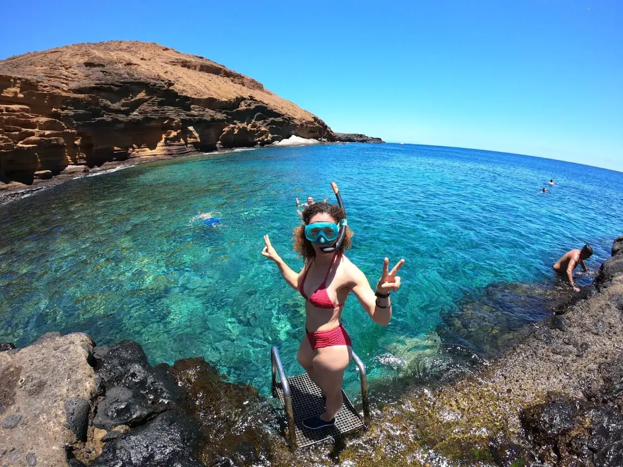 Grudniowe słońce na Wyspach Kanaryjskich. Kobieta w masce do snorkelingu pozdrawia, w tle turkusowa woda i skaliste wybrzeże.
