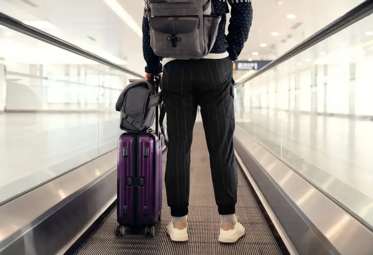 Podr&oacute;żny z fioletową walizką i plecakiem na ruchomych schodach, gotowy do podr&oacute;ży z Qatar Airways. Bagaż podręczny idealnie dopasowany.