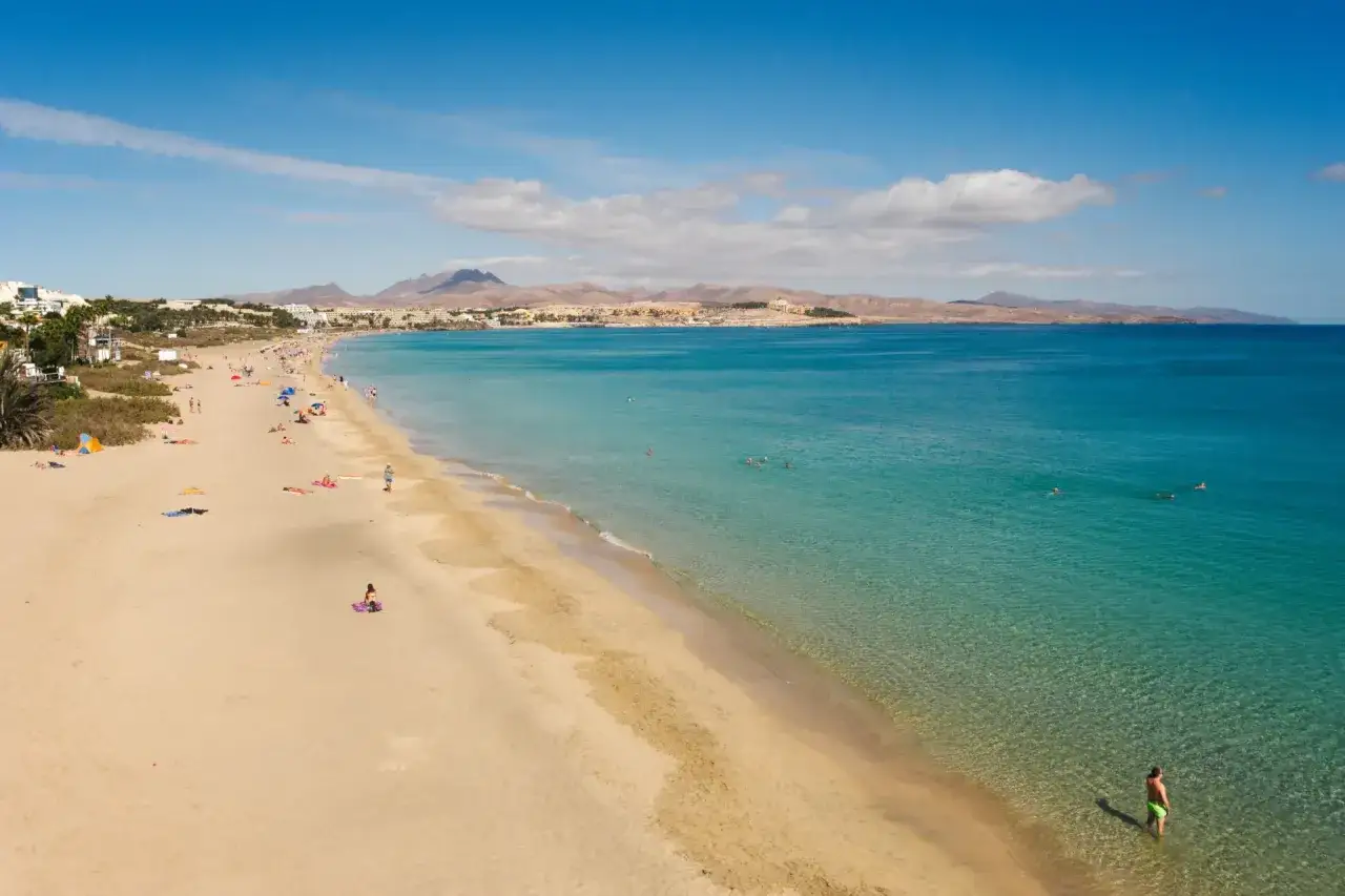 Fuerteventura listopad pogoda: słoneczny dzień na plaży, ludzie opalają się i kąpią w turkusowym morzu.