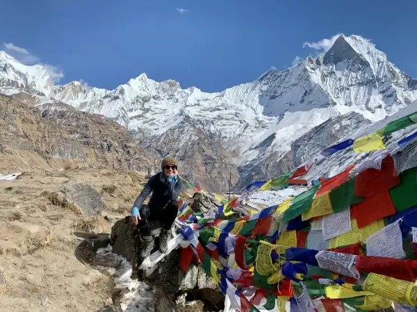 Uśmiechnięta turystka na tle majestatycznych szczytów Annapurna Base Camp, otoczona kolorowymi flagami modlitewnymi.