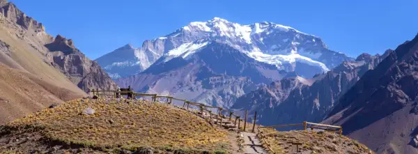 Widok na ośnieżone góry z Aconcagua, z kamienistym szlakiem i drewnianym ogrodzeniem na pierwszym planie.