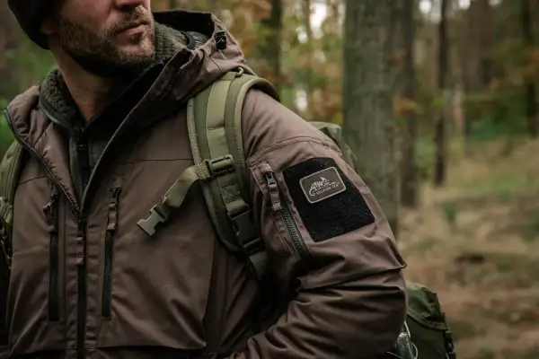 Mężczyzna w brązowej kurtce z naszywką Helikon-Tex, symbol polskie marki outdoorowe, w lesie.